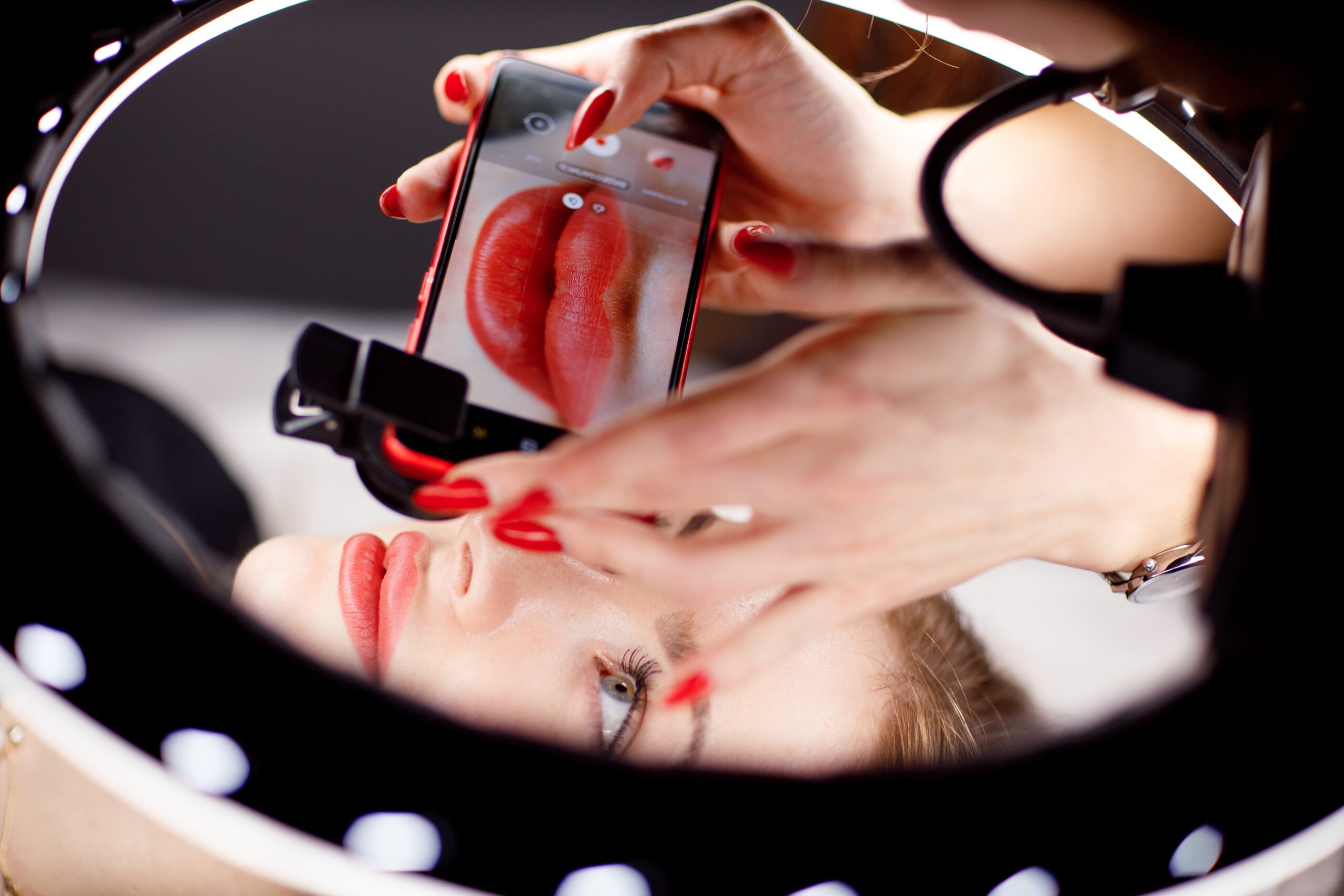 Cosmetologist making photo of work on mobile phone red permanent make up tattoo on young woman lips.